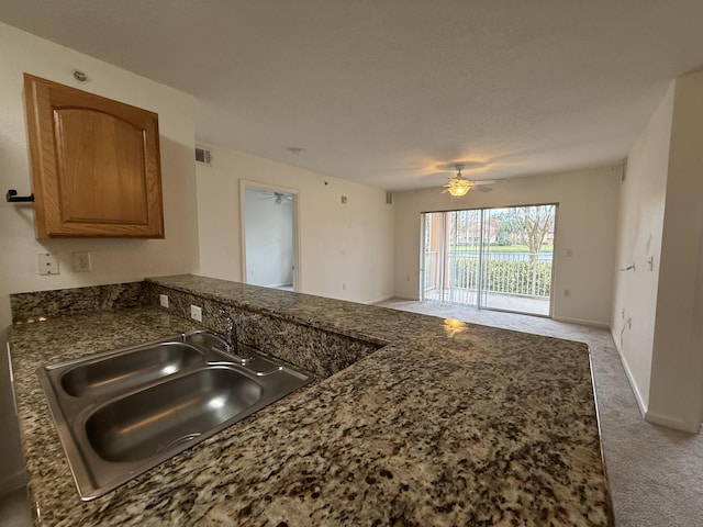 kitchen featuring brown cabinetry, a ceiling fan, a peninsula, light colored carpet, and a sink