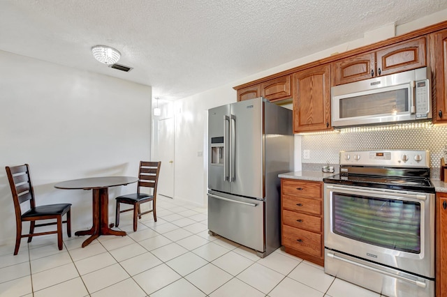 245 Bilbao St, Royal Palm Beach, FL 33411 - kitchen featuring appliances with stainless steel finishes, a textured ceiling, brown cabinetry, decorative backsplash, and light tile patterned floors