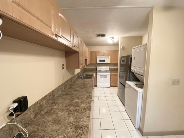 kitchen with white appliances, stacked washing machine and dryer, a sink, dark countertops, and light tile patterned floors