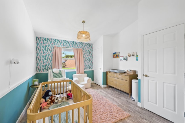 bedroom with wainscoting, a crib, light wood-style floors, wallpapered walls, and baseboards