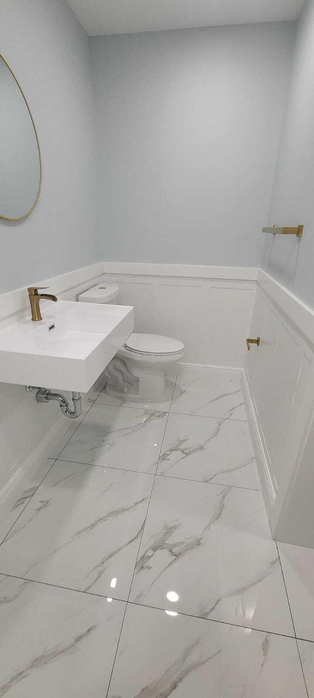 half bath featuring toilet, marble finish floor, a sink, a decorative wall, and a wainscoted wall