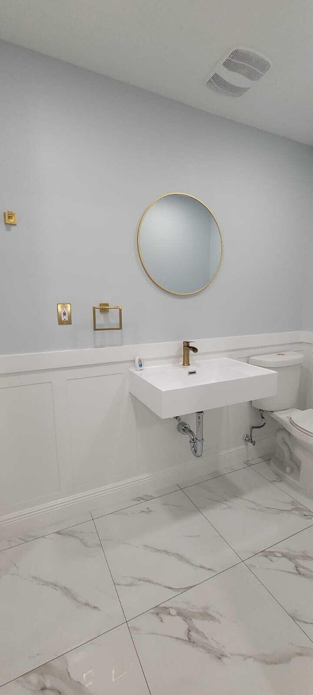 bathroom with toilet, marble finish floor, wainscoting, visible vents, and a sink