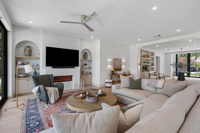 living room featuring built in features, light wood-style flooring, recessed lighting, a warm lit fireplace, and a ceiling fan