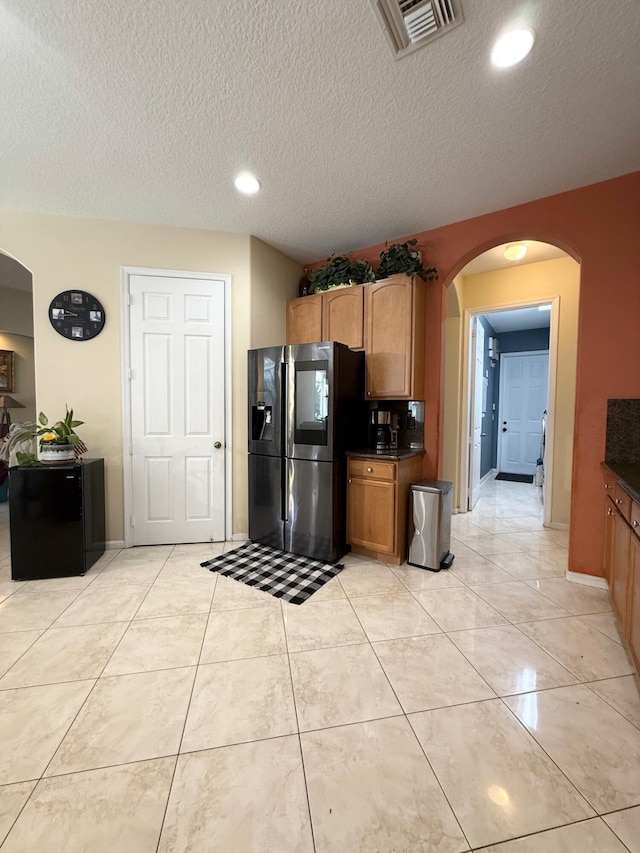 kitchen featuring a textured ceiling, stainless steel refrigerator with ice dispenser, visible vents, arched walkways, and dark countertops
