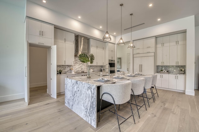 kitchen with light wood finished floors, a kitchen bar, a large island with sink, wall chimney range hood, and decorative backsplash