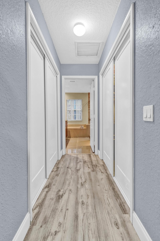 hall with a textured wall, light wood-type flooring, a textured ceiling, and baseboards