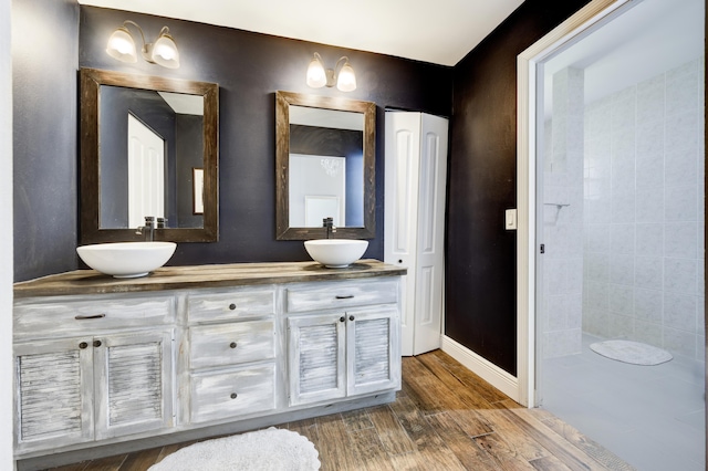 bathroom featuring wood finished floors, double vanity, a tile shower, and baseboards