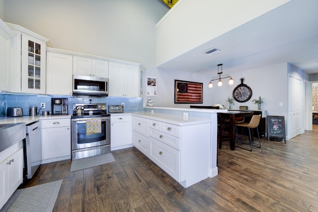 kitchen featuring appliances with stainless steel finishes, a peninsula, dark wood-style flooring, backsplash, and glass insert cabinets