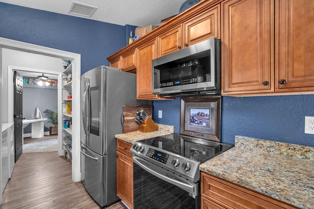 18143 42nd Rd, The Acreage, FL 33470 - kitchen with appliances with stainless steel finishes, a textured wall, brown cabinetry, light wood finished floors, and light stone countertops