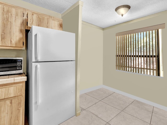 kitchen featuring freestanding refrigerator, tile counters, a textured ceiling, light brown cabinets, and baseboards