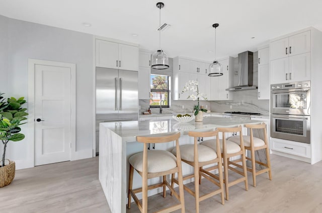 kitchen with wall chimney range hood, stainless steel appliances, light wood-style flooring, a center island, and backsplash