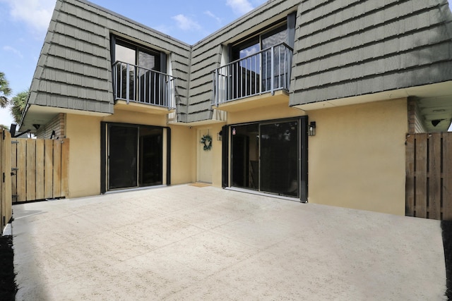 rear view of house with stucco siding, mansard roof, a balcony, a patio, and fence
