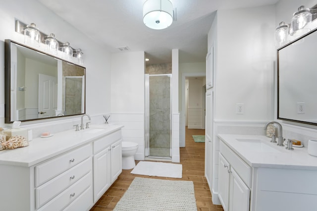 bathroom featuring a stall shower, wood finished floors, toilet, two vanities, and a wainscoted wall