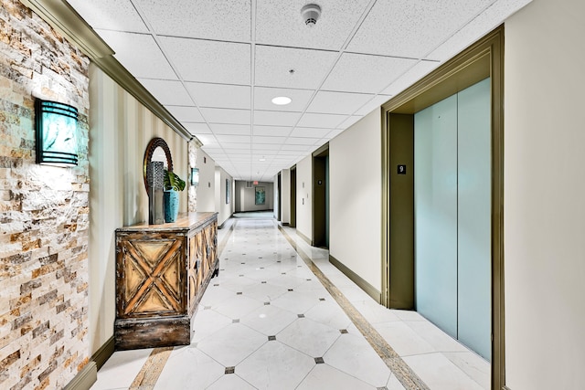 corridor with a paneled ceiling, elevator, inlaid floor details, baseboards, and light tile patterned floors