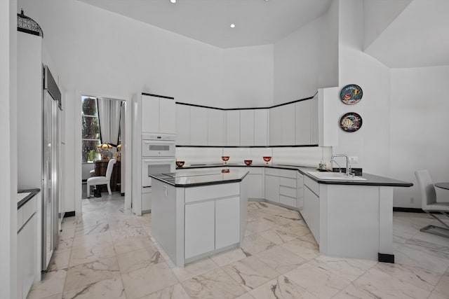 kitchen with a sink, dark countertops, light marble finish flooring, a towering ceiling, and a peninsula