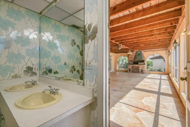 bathroom featuring a sink, wallpapered walls, stone floors, and beamed ceiling