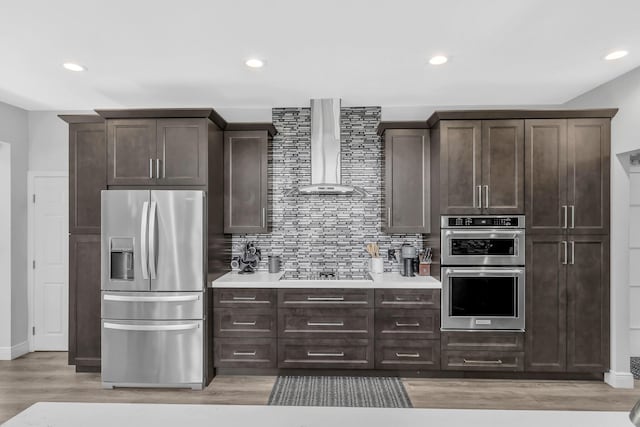 kitchen with stainless steel appliances, wall chimney range hood, light countertops, dark brown cabinetry, and recessed lighting