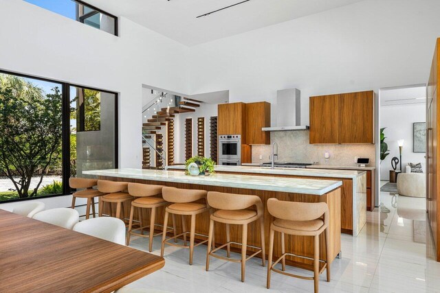 4460 NE 31st Ave, Lighthouse Point, FL 33064 - kitchen featuring wall chimney exhaust hood, double oven, brown cabinets, light countertops, and backsplash