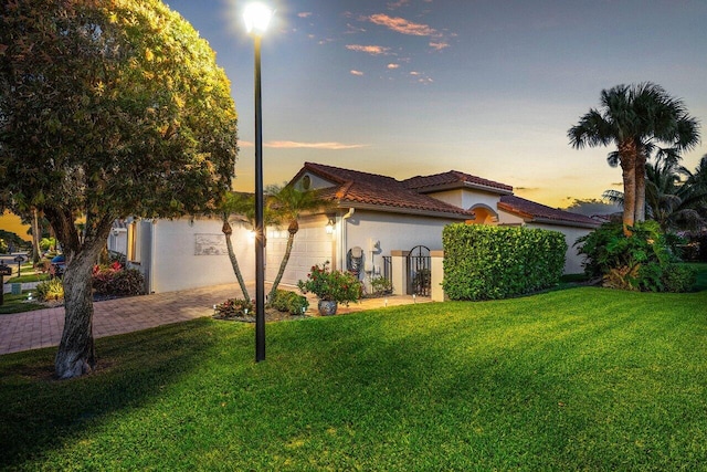 back of property at dusk with stucco siding, a yard, a garage, decorative driveway, and a tiled roof