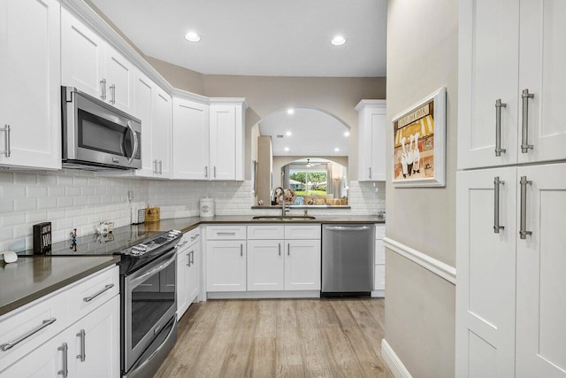 kitchen featuring appliances with stainless steel finishes, a sink, light wood finished floors, dark countertops, and arched walkways