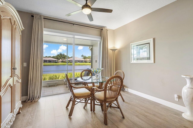dining room with light wood-style floors, a water view, a ceiling fan, baseboards, and a textured ceiling
