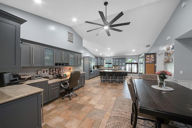kitchen featuring a ceiling fan, high vaulted ceiling, light countertops, glass insert cabinets, and a breakfast bar