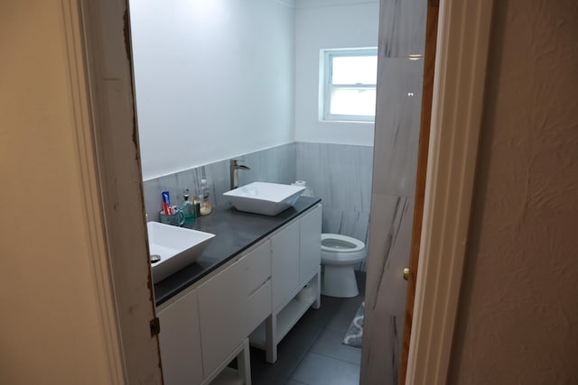 full bath with tile patterned flooring, wainscoting, toilet, double vanity, and tile walls
