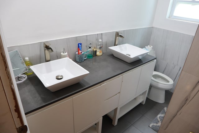 bathroom featuring tile patterned floors, toilet, double vanity, a wainscoted wall, and tile walls
