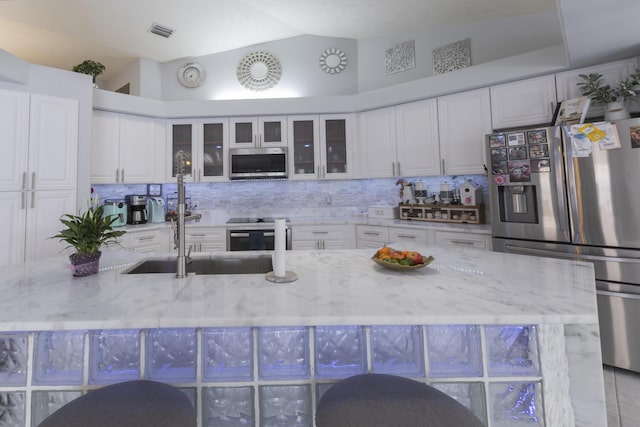 kitchen with appliances with stainless steel finishes, vaulted ceiling, tasteful backsplash, white cabinetry, and glass insert cabinets
