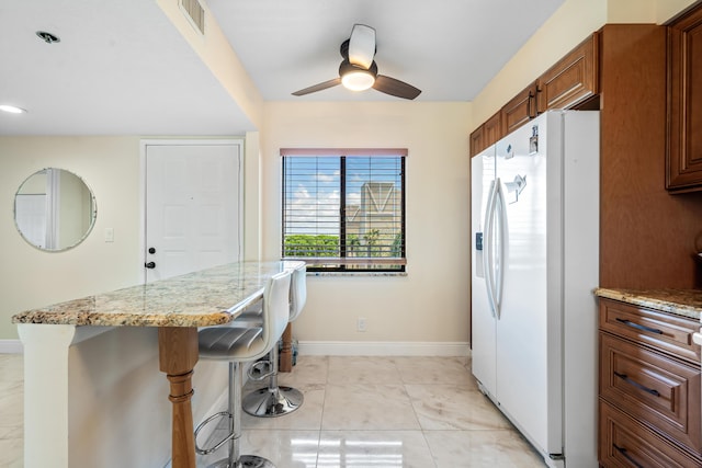 7290 Kinghurst Dr Unit 607, Delray Beach, FL 33446 - kitchen with white refrigerator with ice dispenser, a kitchen bar, brown cabinetry, ceiling fan, and light stone counters