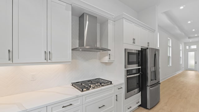 kitchen featuring appliances with stainless steel finishes, wall chimney range hood, white cabinets, light stone counters, and backsplash