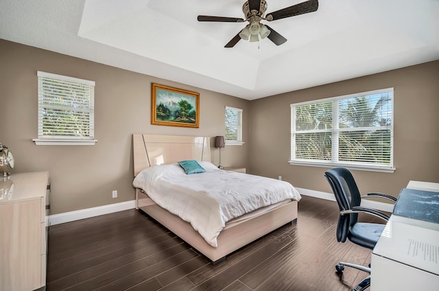 bedroom featuring a raised ceiling, dark wood-type flooring, baseboards, ceiling fan, and a desk