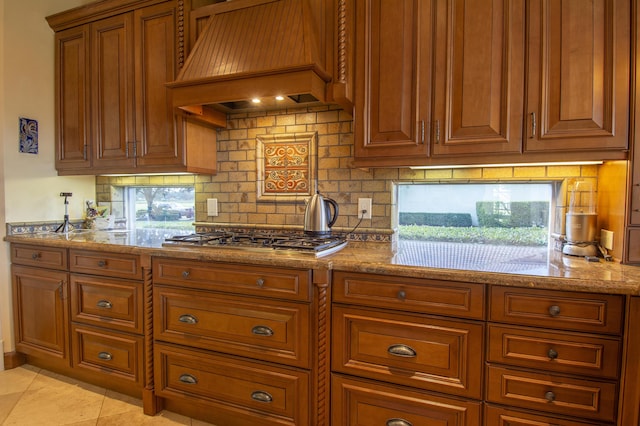 143 SE Bella Strano, Port Saint Lucie, FL 34984 - kitchen featuring stainless steel gas cooktop, custom range hood, brown cabinets, light stone countertops, and backsplash