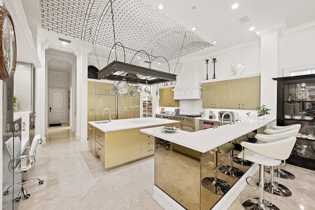 kitchen featuring ornate columns, a towering ceiling, crown molding, a kitchen island with sink, and a breakfast bar