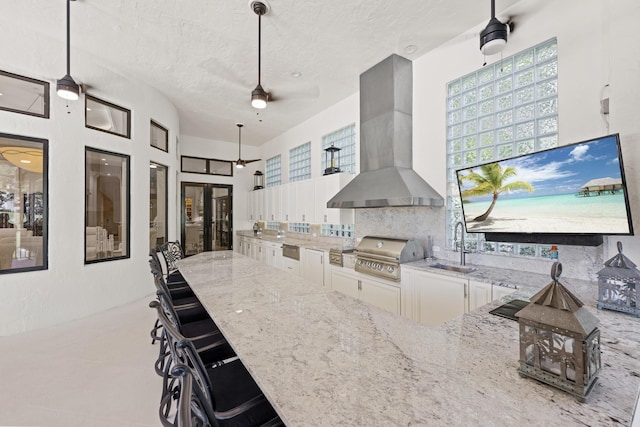 kitchen featuring wall chimney exhaust hood, light stone counters, a ceiling fan, white cabinetry, and tasteful backsplash