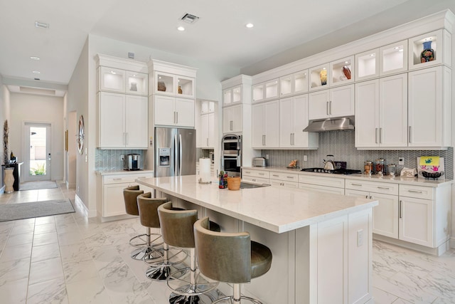 kitchen featuring stainless steel appliances, under cabinet range hood, light marble finish floors, an island with sink, and recessed lighting
