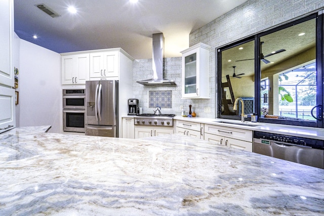 kitchen with wall chimney range hood, appliances with stainless steel finishes, a sink, glass insert cabinets, and light stone countertops
