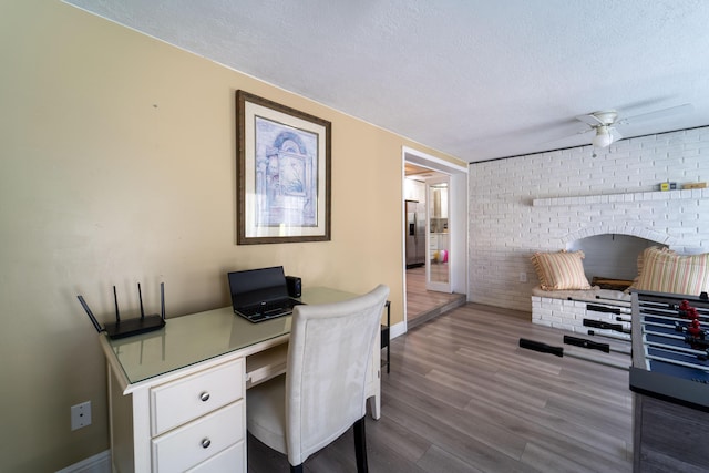 office area featuring a textured ceiling, wood finished floors, a ceiling fan, brick wall, and baseboards