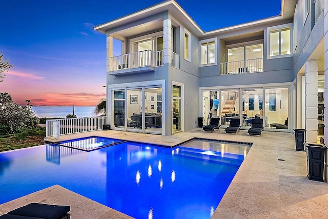 back of house at dusk featuring a pool with connected hot tub, a balcony, a patio, and a water view