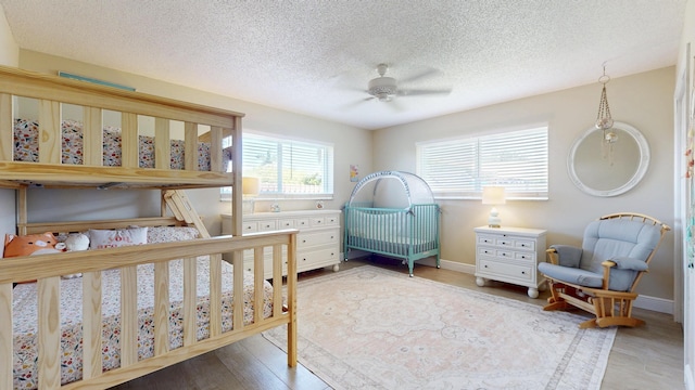 bedroom featuring a textured ceiling, wood finished floors, a crib, baseboards, and a ceiling fan