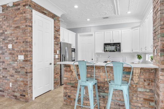 1909 Flower Dr, Palm Beach Gardens, FL 33410 - kitchen with black microwave, light tile patterned floors, a breakfast bar, white cabinets, and brick wall