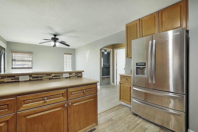 kitchen featuring stainless steel refrigerator with ice dispenser, arched walkways, a ceiling fan, brown cabinets, and light wood-style floors