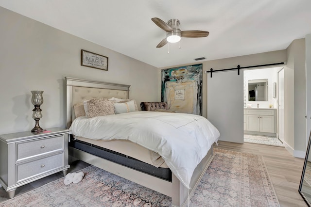 bedroom with a barn door, light wood-type flooring, ensuite bath, and ceiling fan