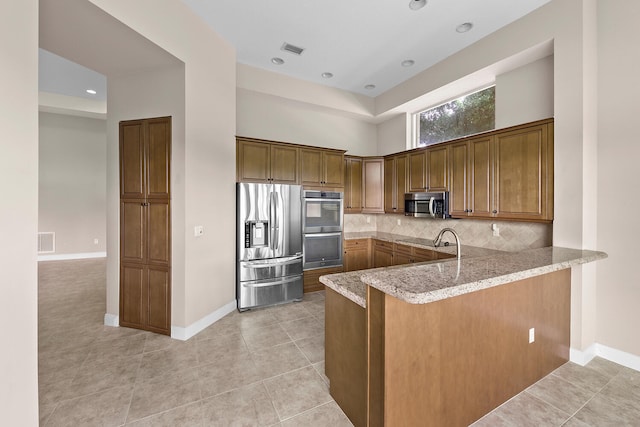9724 Via Grandezza, Wellington, FL 33411 - kitchen featuring stainless steel appliances, a peninsula, light stone counters, backsplash, and brown cabinetry