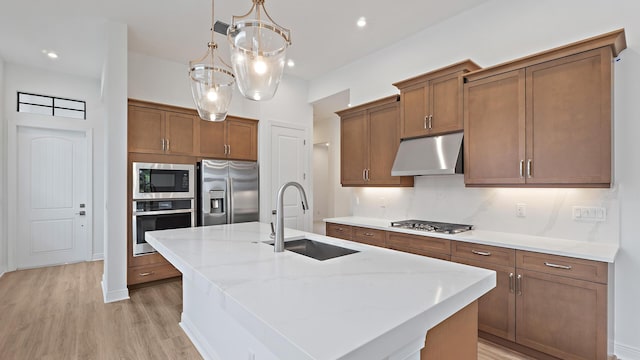 203 SE Via Sangro, Port Saint Lucie, FL 34952 - kitchen featuring appliances with stainless steel finishes, under cabinet range hood, a kitchen island with sink, brown cabinetry, and light wood-type flooring