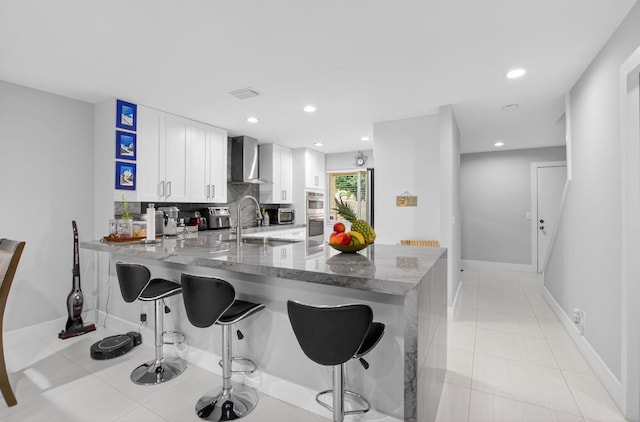 3469 Deer Creek Palladian Cir, Deerfield Beach, FL 33442 - kitchen featuring wall chimney exhaust hood, a peninsula, white cabinetry, light stone countertops, and a kitchen bar