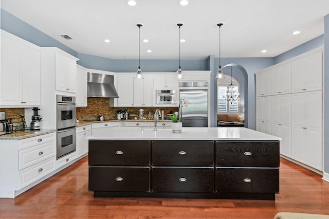 908 SW Squire Johns Ln, Palm City, FL 34990 - kitchen featuring appliances with stainless steel finishes, wall chimney range hood, a kitchen island with sink, tasteful backsplash, and white cabinetry