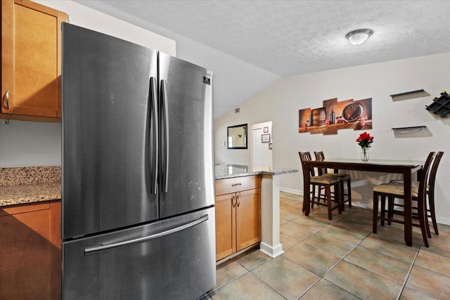6015 Raintree Trl, Fort Pierce, FL 34982 - kitchen featuring freestanding refrigerator, brown cabinetry, light tile patterned floors, lofted ceiling, and light stone countertops