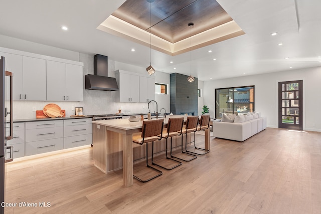 kitchen featuring white cabinets, wall chimney exhaust hood, pendant lighting, light hardwood / wood-style flooring, and a kitchen island with sink