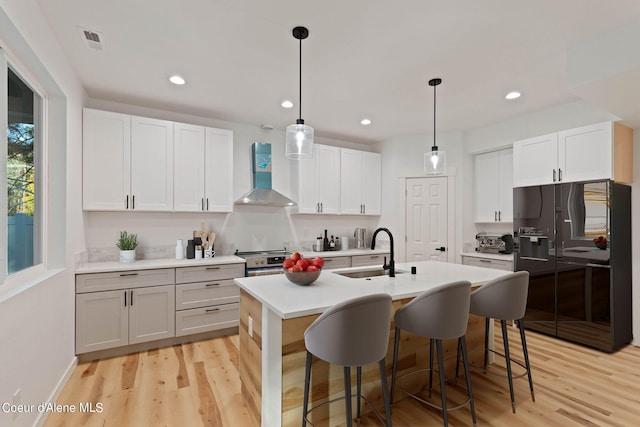 kitchen with wall chimney range hood, light wood-type flooring, black fridge, and a center island with sink